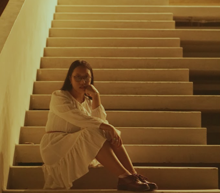 Indonesian musical artist Shelma Shalindri dressed in white sitting at the bottom of a staircase with a soft glowing light at the top of the staircase