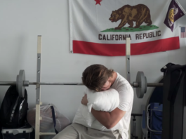 Ryan Beatty clutching a white pillow and burying his face in it while sitting on a weightlifting bench with the California state flag behind him in the "Ribbons" music video