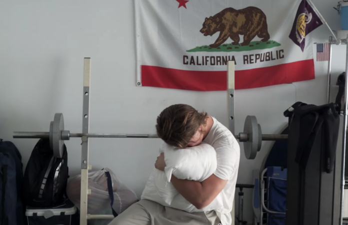 Ryan Beatty clutching a white pillow and burying his face in it while sitting on a weightlifting bench with the California state flag behind him in the 